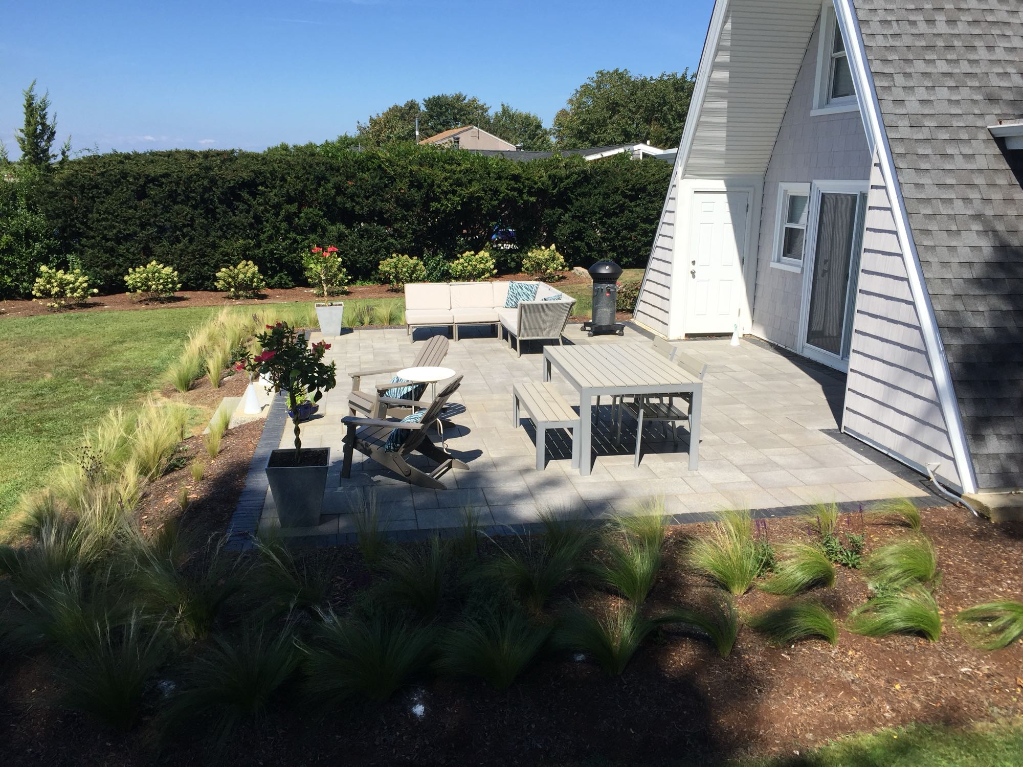 Sapowet patio project; side view of A-frame house with small paver patio and natural stone steps with nice patio furniture and hedges in background