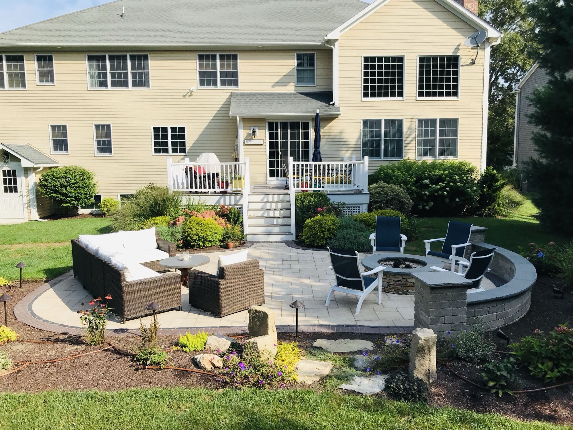 Sylvan Court project; view of paver patio with short wall around fire pit area and section couch area surrounded by beautiful landscaping with large, two-story house in the background