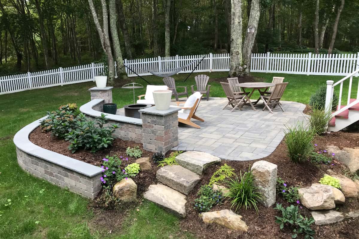 small stone patio with multi-level landscaping in a white picket fenced backyard