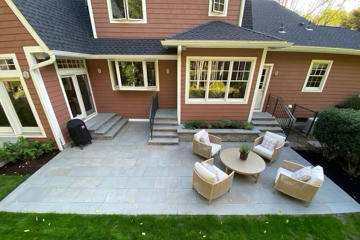 A large outdoor living space with light gray stone pavers, furnished with four wicker chairs around a coffee table, connecting to a reddish-brown house via stone steps.