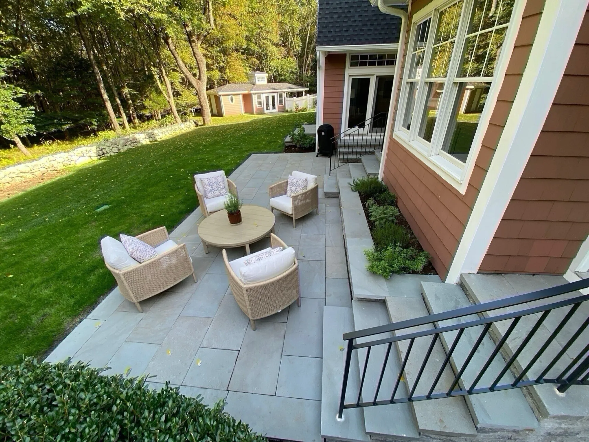 A professionally installed stone patio uses large gray pavers and includes built-in stone steps with a black railing leading to a home. Wicker furniture sits on the terrace overlooking a green lawn and forest.