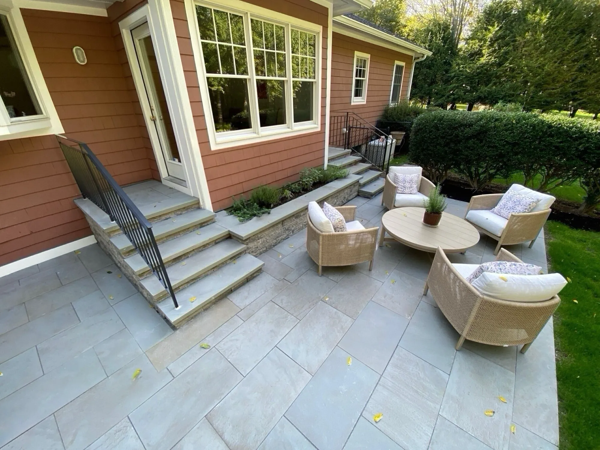 An outdoor living space featuring a smooth, grey flagstone patio and matching stone steps leading down from the house. Four wicker chairs surround a low table.