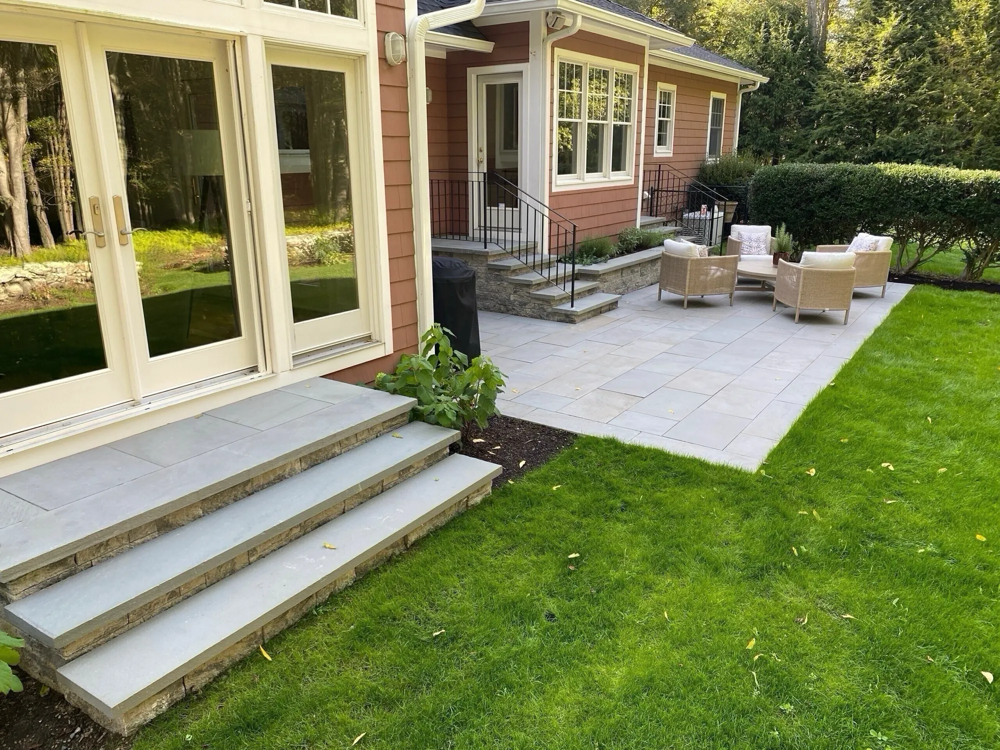 A modern stone patio and wide blue-gray stone steps lead from a house's French doors to a vibrant green lawn. Wicker seating surrounds a table on the patio.